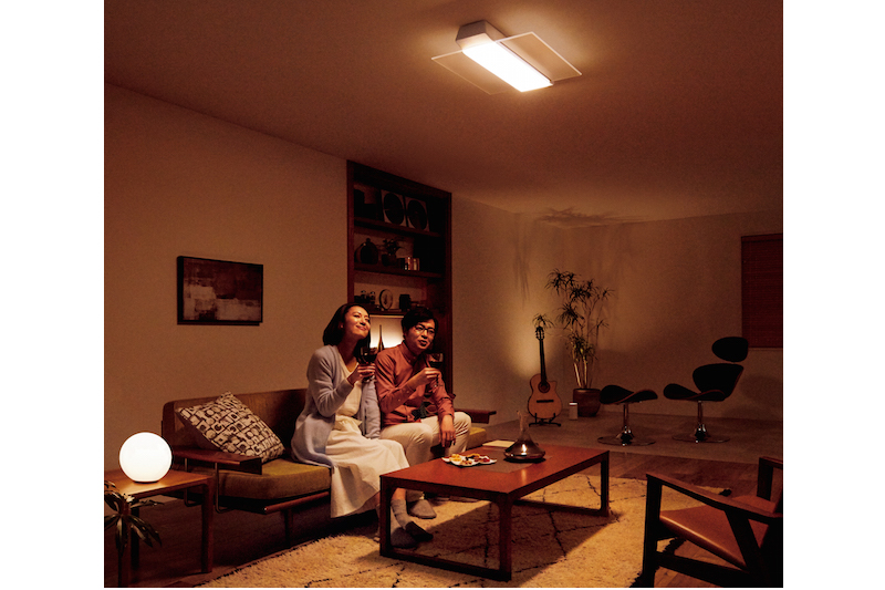 パナソニック『AIR PANEL LED THE SOUND HH-XCF1203A（LINK STYLE LED対応）』のLED照明がついているリビングのソファに腰掛けワインを飲んでいる男性と女性