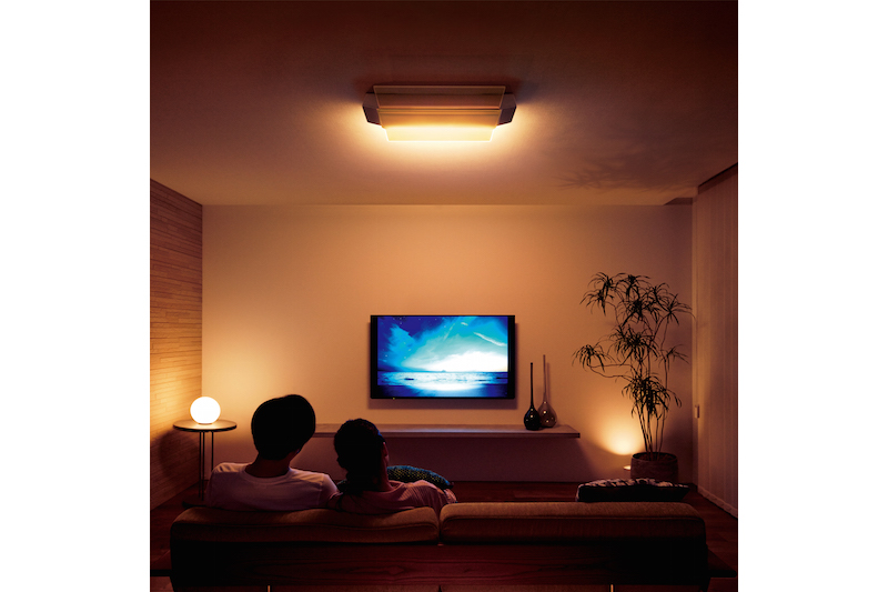 パナソニック『AIR PANEL LED THE SOUND HH-XCF1203A（LINK STYLE LED対応）』のLED照明が暗めについているリビングでソファに腰掛けテレビを観ている男性と女性