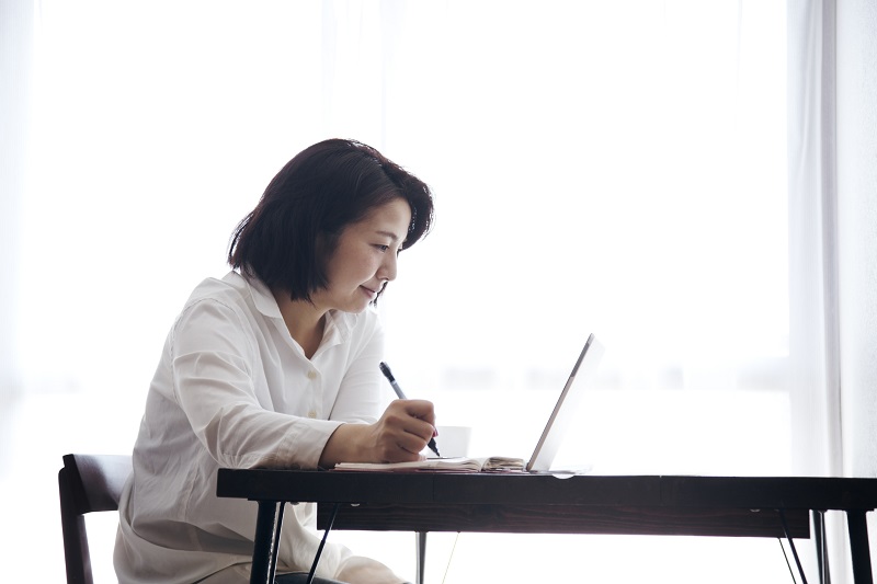 テーブルの上のノートパソコンを見ながら紙に書き込む女性