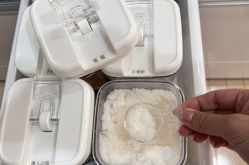 キッチンの下の引き出しの収納に入れられた蓋が白で本体が透明の容器のうち、ひとつの蓋開け、中の粗塩を計量スプーンですくっている手元