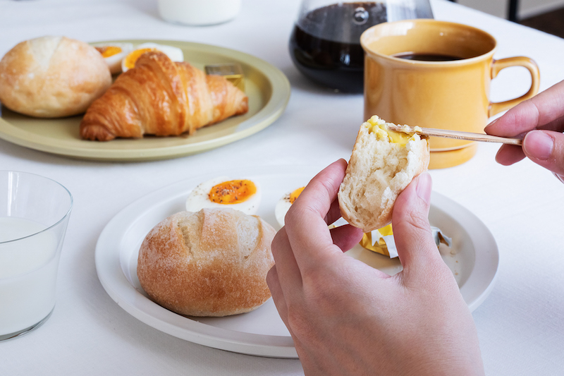 テーブルにクロワッサンとテーブルパンが乗った皿や珈琲があり、パンにバターを塗っている手元