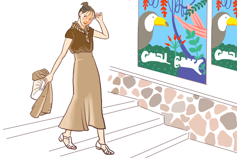 柄が飾られている壁の横の階段を降りる、茶色のTシャツにロングスカートを着た女性