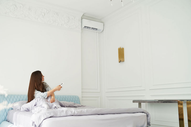 ベッドの上に座っている女性が、リモコンを片手に、部屋の上部に設置されたエアコンを操作している